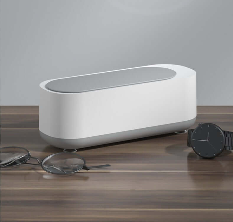 Ultrasonic jewelry cleaner on a wooden table beside glasses and a watch, designed for effective cleaning.